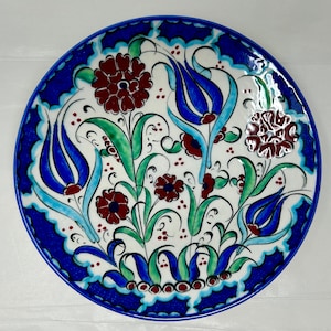 May include: A white ceramic plate with a blue rim and a hand-painted design of blue and green flowers with red accents.