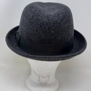 Pode incluir: Um chap&eacute;u fedora de feltro cinza-escuro com uma copa estruturada e aba ligeiramente curva. O chap&eacute;u &eacute; acentuado com uma fita de gorgor&atilde;o preta s&oacute;lida que circunda a base da copa, proporcionando um contraste distinto com o feltro cinza texturizado.
