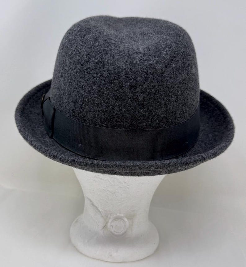 Pode incluir: Um chap&eacute;u fedora de feltro cinza-escuro com uma copa estruturada e aba ligeiramente curva. O chap&eacute;u &eacute; acentuado com uma fita de gorgor&atilde;o preta s&oacute;lida que circunda a base da copa, proporcionando um contraste distinto com o feltro cinza texturizado.