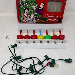 Può includere: Set di luci a bolle Holiday Lane vintage con una scatola rossa raffigurante una famiglia che decora un albero di Natale. Il set include sette luci a bolle colorate in rosso, giallo e verde, montate su un espositore bianco. È incluso anche un cavo elettrico verde.