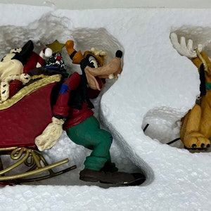 May include: A set of three Christmas ornaments featuring Mickey Mouse, Goofy, and Pluto. Mickey is in a red sleigh with a gold trim, Goofy is holding a red bag, and Pluto is standing with his antlers up.