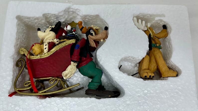 May include: A set of three Christmas ornaments featuring Mickey Mouse, Goofy, and Pluto. Mickey is in a red sleigh with a gold trim, Goofy is holding a red bag, and Pluto is standing with his antlers up.