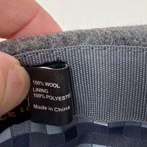 Pode incluir: O interior de um chap&eacute;u de l&atilde; cinzenta com uma etiqueta de tecido preta com texto branco que diz 100% WOOL, LINING: 100% POLYESTER e Made in China. O chap&eacute;u tem uma banda interior cinzenta texturizada e um forro com padr&atilde;o xadrez azul e cinzento.