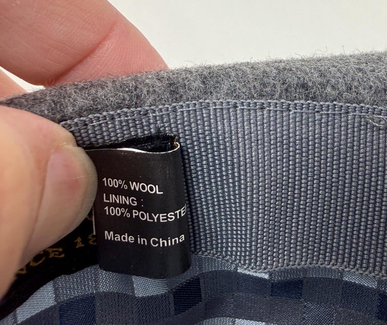 Pode incluir: O interior de um chap&eacute;u de l&atilde; cinzenta com uma etiqueta de tecido preta com texto branco que diz 100% WOOL, LINING: 100% POLYESTER e Made in China. O chap&eacute;u tem uma banda interior cinzenta texturizada e um forro com padr&atilde;o xadrez azul e cinzento.