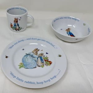 Puede incluir: Un juego de vajilla infantil de cerámica blanca con ilustraciones de Peter Rabbit. El juego incluye una taza, un cuenco y un plato. El plato tiene el texto "Hop little rabbit, hop hop hop" y "Now run along...and don't get into mischief."