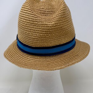 May include: A tan straw fedora hat featuring a pinched crown and a wide brim. The hat is accented with a decorative band consisting of a bright blue center stripe bordered by two darker navy blue stripes encircling the base of the crown.