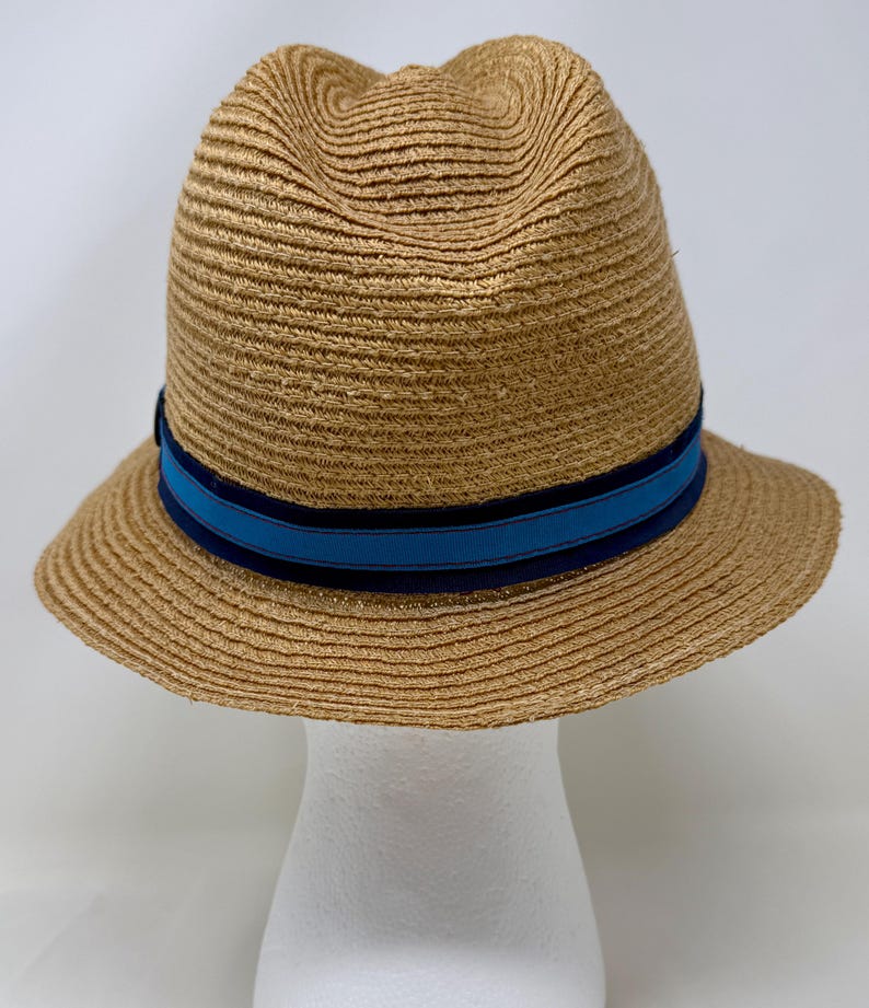May include: A tan straw fedora hat featuring a pinched crown and a wide brim. The hat is accented with a decorative band consisting of a bright blue center stripe bordered by two darker navy blue stripes encircling the base of the crown.