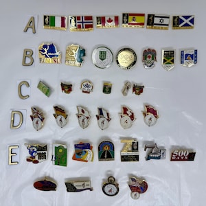May include: A selection of collectible pins showcasing flags, Olympic symbols, and commemorative designs. The pins include flags from different nations, Olympic rings, and pins with text such as "Atlanta 1996" and "500 Days".
