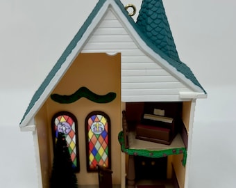 May include: A miniature church ornament with a green roof, white siding, and stained glass windows. The interior features a small Christmas tree, a wooden pew, and an organ.
