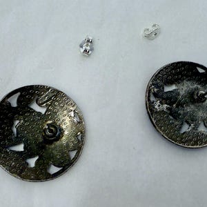 May include: A pair of silver-toned, round stud earrings with a floral design and cut-out details. The earrings have a textured surface and are accompanied by two silver earring backs. The earrings are displayed on a white surface.