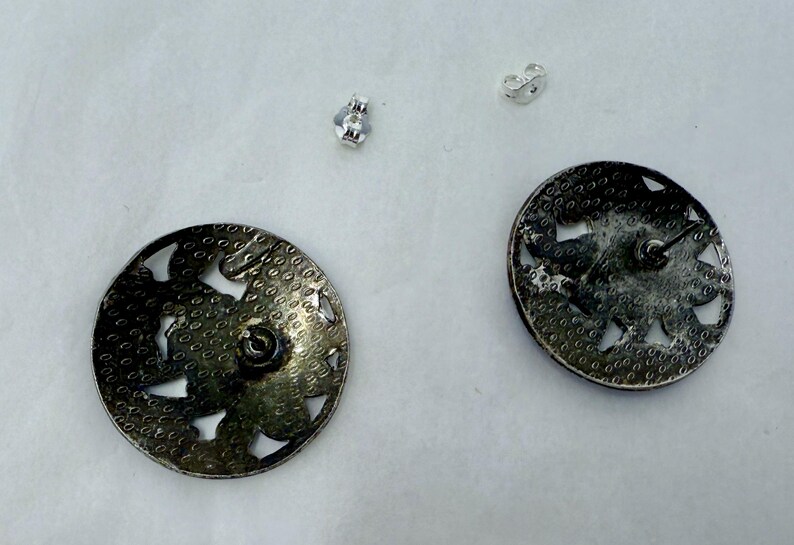 May include: A pair of silver-toned, round stud earrings with a floral design and cut-out details. The earrings have a textured surface and are accompanied by two silver earring backs. The earrings are displayed on a white surface.