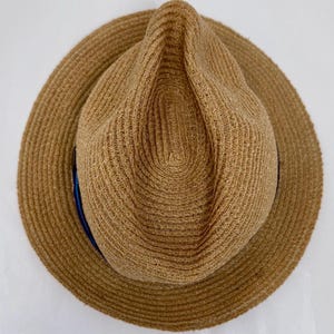 May include: A tan straw fedora hat with a pinched crown and a wide brim. The hat features a woven texture throughout the material and a thin, dark blue ribbon band encircling the base of the crown, visible on the left side of the hat.