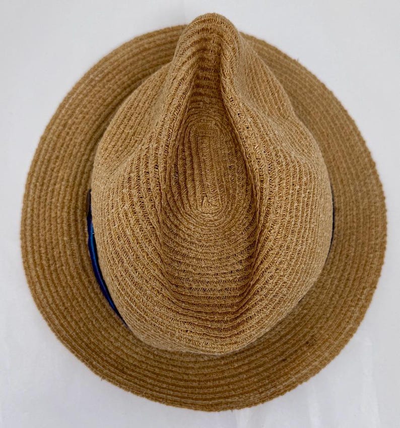 May include: A tan straw fedora hat with a pinched crown and a wide brim. The hat features a woven texture throughout the material and a thin, dark blue ribbon band encircling the base of the crown, visible on the left side of the hat.