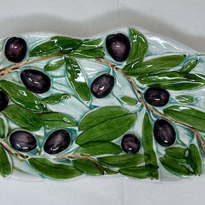 Puede incluir: Una fuente de cerámica para servir aceitunas con forma de rama de olivo. Presenta hojas verdes, tallos marrones y aceitunas moradas oscuras. El fondo es de color azul claro.