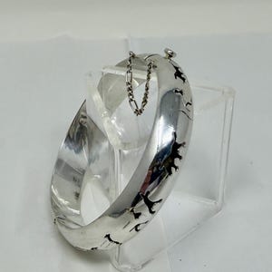 Op de afbeelding: Een brede, zilverkleurige armband met uitgesneden dierensilhouetten. De armband heeft een scharnierende sluiting en een veiligheidsketting. Het ontwerp toont een reeks gestileerde dierenfiguren rond de band. De armband wordt gepresenteerd op een helder, schuin oppervlak.