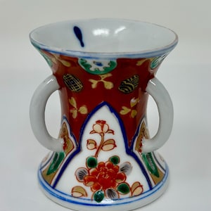 May include: A small, white porcelain vase with two handles. The vase is decorated with a red, green, and gold floral pattern.