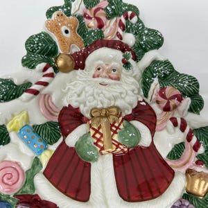 May include: A ceramic Christmas tree-shaped serving dish features a Santa Claus figure holding a gift, surrounded by candy canes, lollipops, and other festive decorations. The dish is white with green, red, and gold accents.