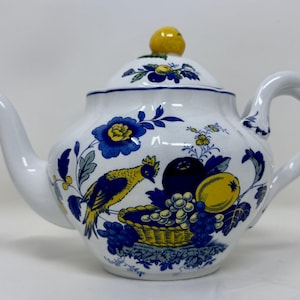 Puede incluir: Tetera de cerámica blanca con tapa, decorada con motivos florales y frutales azules y amarillos. El diseño incluye un pájaro, uvas y una cesta. El asa y el pico son blancos, y la tapa tiene un pomo amarillo.