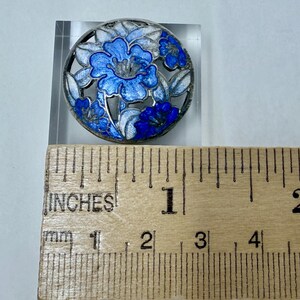 May include: Round, silver-toned brooch with a floral design. The brooch features blue and white enamel flowers within a circular frame. The brooch measures approximately 1 inch in diameter.