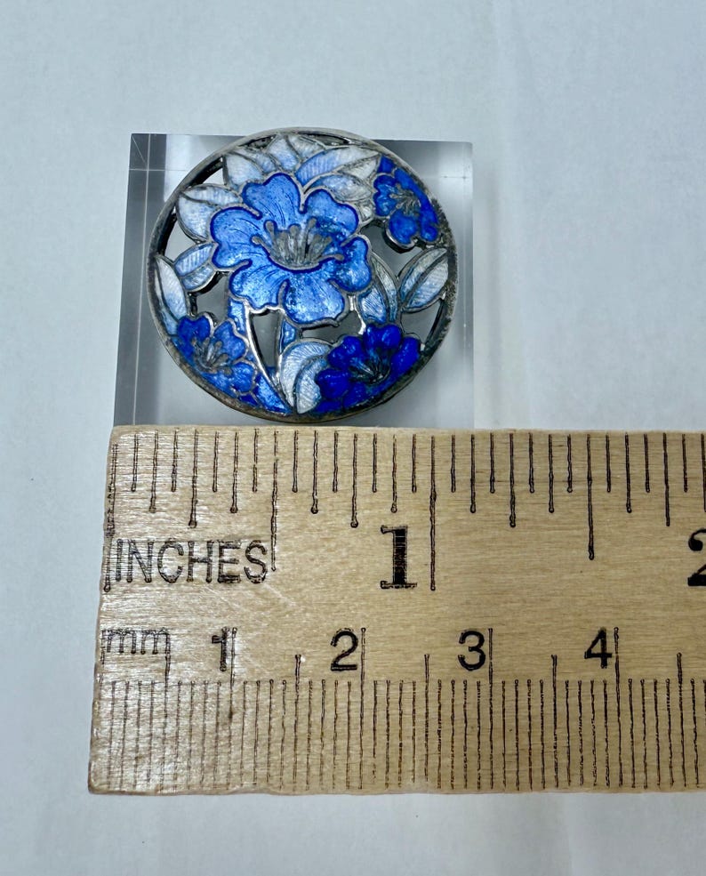 May include: Round, silver-toned brooch with a floral design. The brooch features blue and white enamel flowers within a circular frame. The brooch measures approximately 1 inch in diameter.