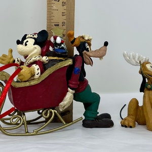 May include: A set of three Christmas figurines featuring Mickey Mouse, Goofy, and Pluto. Mickey is dressed as Santa Claus and riding in a red sleigh. Goofy is wearing a green and blue outfit and is pushing the sleigh. Pluto is wearing a reindeer antler headband and is sitting on the ground.
