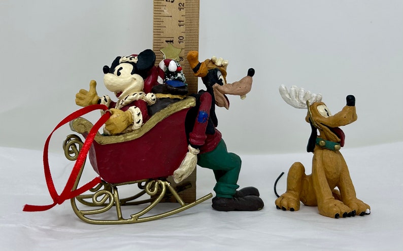 May include: A set of three Christmas figurines featuring Mickey Mouse, Goofy, and Pluto. Mickey is dressed as Santa Claus and riding in a red sleigh. Goofy is wearing a green and blue outfit and is pushing the sleigh. Pluto is wearing a reindeer antler headband and is sitting on the ground.