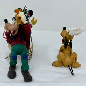 May include: Two Christmas ornaments featuring Goofy and Pluto. Goofy is dressed in a red and green outfit and is pulling a sled with a small Christmas tree. Pluto is dressed in a red and green outfit and has antlers. Both ornaments are made of painted ceramic and have a wire hanger.