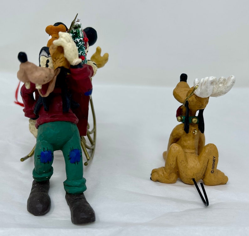 May include: Two Christmas ornaments featuring Goofy and Pluto. Goofy is dressed in a red and green outfit and is pulling a sled with a small Christmas tree. Pluto is dressed in a red and green outfit and has antlers. Both ornaments are made of painted ceramic and have a wire hanger.