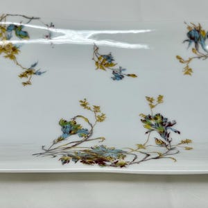 Puede incluir: Un plato rectangular blanco para servir con un borde festoneado. El plato está decorado con un patrón floral en tonos azules, verdes y amarillos.