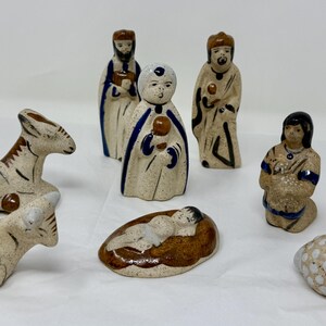 May include: A ceramic nativity set featuring figures of the Holy Family, the Three Wise Men, and animals. The figures are painted in earth tones with blue and brown accents. The set includes a baby Jesus in a manger and two sheep.