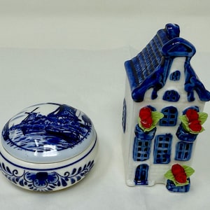 May include: Two ceramic decorative pieces: a round trinket box with a windmill scene and a house-shaped figurine. Both are white with blue accents and floral patterns. The house has red flower accents.