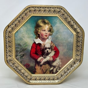 Puede incluir: Caja de hojalata octogonal con un borde dorado adornado. El centro de la caja presenta una pintura de un niño pequeño con una chaqueta roja sosteniendo un perro blanco.