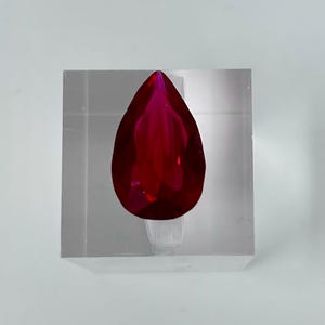 Puede incluir: Una piedra preciosa de color rojo intenso en forma de pera, expuesta en un soporte de acrílico transparente.