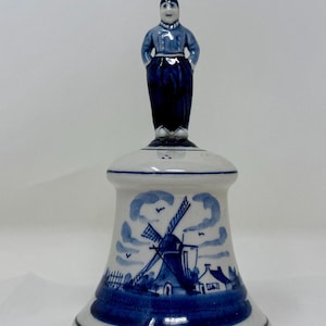 May include: A ceramic bell with a Dutch theme. The bell is white with blue accents and features a windmill scene and a figure of a person in traditional Dutch attire. The bell is approximately 6 inches tall.