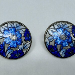 May include: A pair of round earrings with a floral design. The earrings feature a silver-colored metal frame with a cutout pattern. The center of each earring has a blue flower with white and blue leaves. The earrings are approximately 1 inch in diameter.