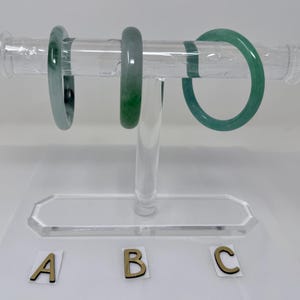 May include: Three jade bangles in varying shades of green are displayed on a clear acrylic jewelry stand. The bangles are labeled A, B, and C with gold-colored letters. The stand has a rectangular base and a cylindrical support.