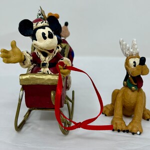 May include: A Christmas-themed figurine set featuring Mickey Mouse dressed as Santa Claus in a red sleigh pulled by Pluto with reindeer antlers. Mickey is waving and Pluto is looking forward with his tongue sticking out.