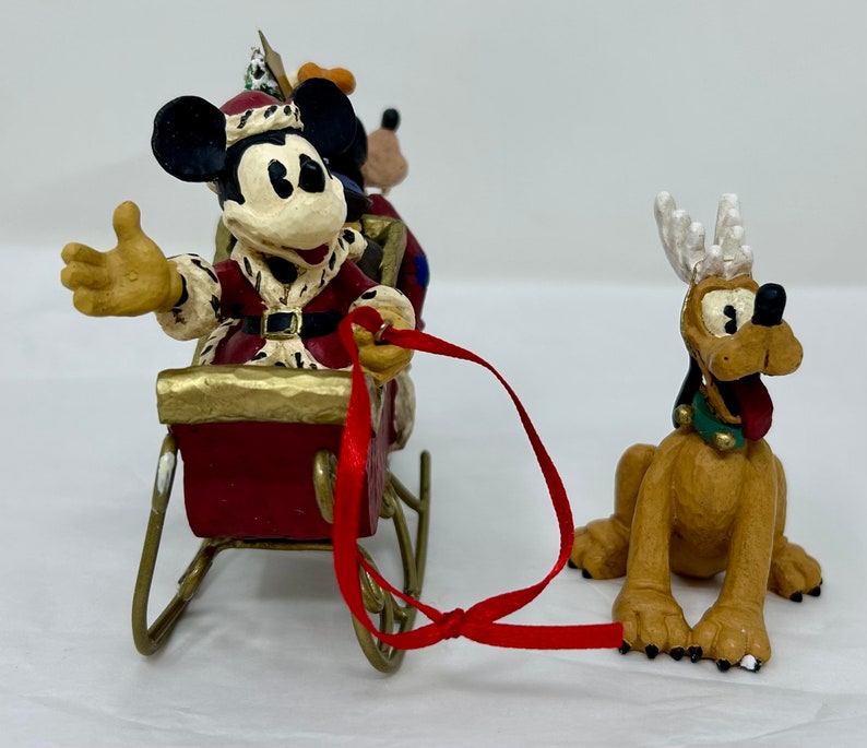 May include: A Christmas-themed figurine set featuring Mickey Mouse dressed as Santa Claus in a red sleigh pulled by Pluto with reindeer antlers. Mickey is waving and Pluto is looking forward with his tongue sticking out.
