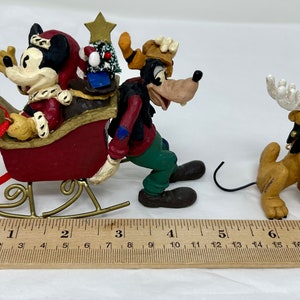 May include: A Christmas ornament featuring Mickey Mouse, Goofy, and Pluto. Mickey is dressed as Santa Claus and riding in a red sleigh pulled by Goofy. Pluto is wearing a reindeer antler headband and is standing behind Goofy.