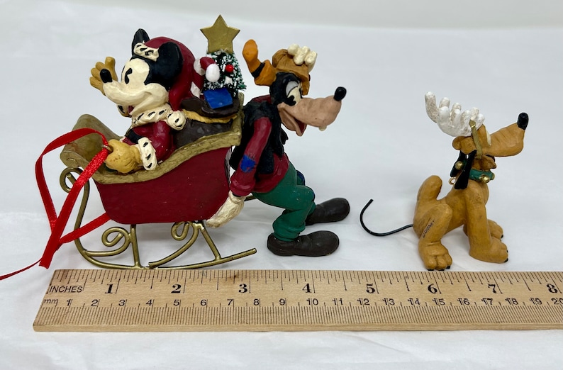 May include: A Christmas ornament featuring Mickey Mouse, Goofy, and Pluto. Mickey is dressed as Santa Claus and riding in a red sleigh pulled by Goofy. Pluto is wearing a reindeer antler headband and is standing behind Goofy.