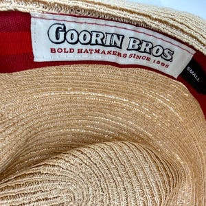 May include: The interior of a tan straw hat featuring a white fabric label with the black text Goorin Bros. Bold Hatmakers Since 1895. A small black tag indicates the size as Small. The inner lining is a vibrant red fabric with a subtle stripe pattern.