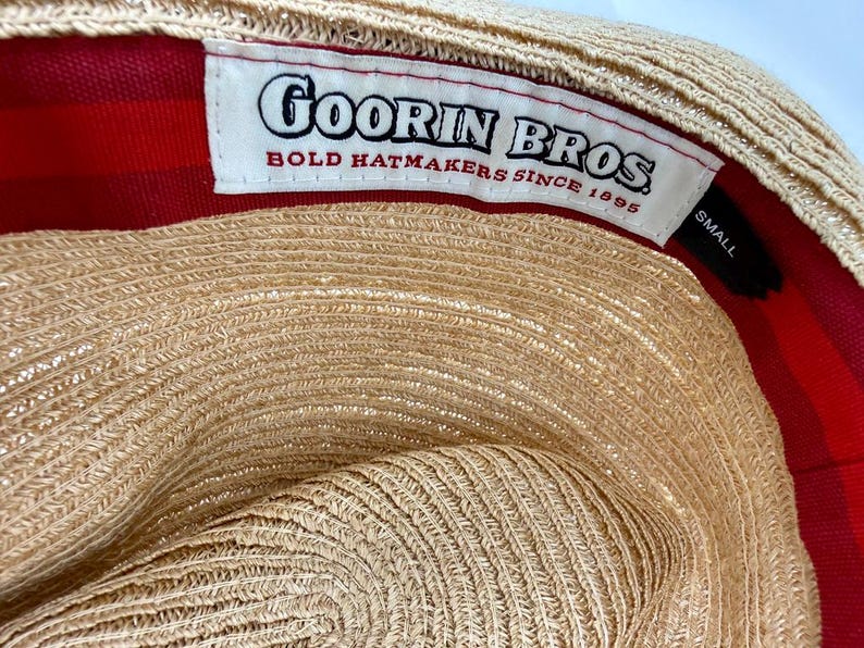 May include: The interior of a tan straw hat featuring a white fabric label with the black text Goorin Bros. Bold Hatmakers Since 1895. A small black tag indicates the size as Small. The inner lining is a vibrant red fabric with a subtle stripe pattern.