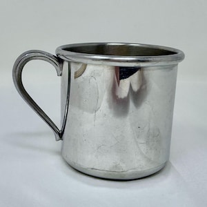 May include: A small, silver-colored metal mug with a curved handle. The mug has a cylindrical body and a slightly flared rim. The handle is attached to the side of the mug and is curved in a simple, elegant design. The mug appears to be made of stainless steel.