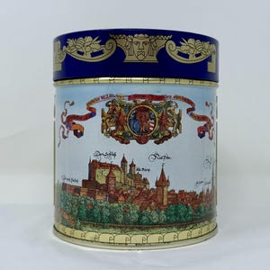 May include: A cylindrical tin with a blue lid and a light blue body. The lid has a gold decorative pattern. The body features a detailed illustration of a European town with a castle, trees, and text in German.