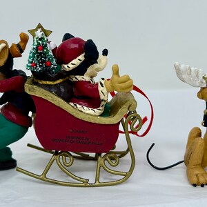 May include: A Christmas ornament featuring Mickey Mouse, Goofy, and Pluto. Mickey is dressed as Santa Claus and is riding in a red sleigh pulled by Goofy. Pluto is dressed as a reindeer and is standing behind the sleigh. The ornament is made of ceramic and is decorated with festive details.