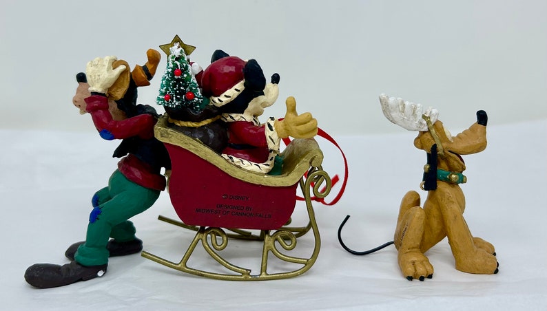May include: A Christmas ornament featuring Mickey Mouse, Goofy, and Pluto. Mickey is dressed as Santa Claus and is riding in a red sleigh pulled by Goofy. Pluto is dressed as a reindeer and is standing behind the sleigh. The ornament is made of ceramic and is decorated with festive details.