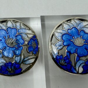 May include: Two round, silver-toned earrings with a floral design. Each earring features a blue flower with white and blue leaves, set within a circular, openwork frame. The earrings are displayed on clear acrylic stands.