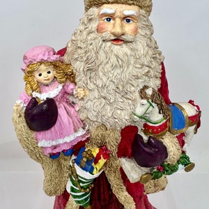 May include: A Santa Claus figurine holding a doll and a rocking horse. He is wearing a red suit with white fur trim and a white beard. The doll is wearing a pink dress and the rocking horse is white with blue and gold accents.