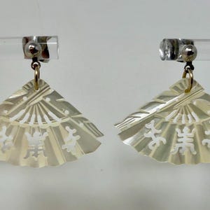 May include: A pair of fan-shaped earrings. The earrings are made of a pearlescent material with a carved design. The design includes a fan shape with intricate lines and symbols. The earrings are attached to silver-colored clips.