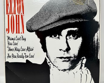 May include: Album cover featuring Elton John in a black and white portrait, wearing a patterned cap. The artist's name is in red, with song titles like "Mama Can't Buy You Love" listed below. The album is a vintage music collectible.